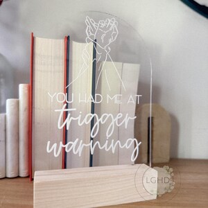 May include: Clear acrylic sign with the text "YOU HAD ME AT trigger warning" in white script. The sign features a line drawing of two hands clasped together. The sign is displayed on a wooden base.