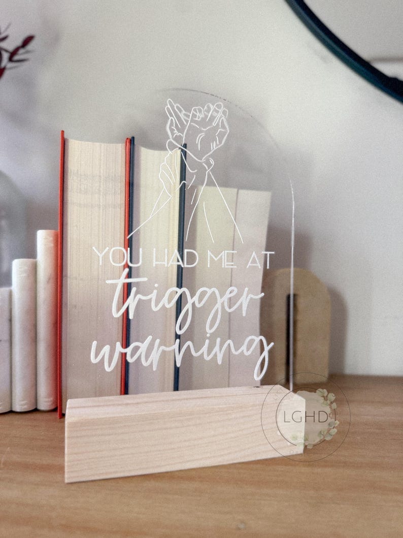 May include: Clear acrylic sign with the text "YOU HAD ME AT trigger warning" in white script. The sign features a line drawing of two hands clasped together. The sign is displayed on a wooden base.