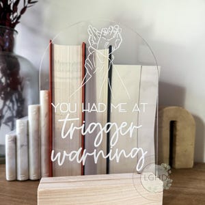 May include: Clear acrylic sign with the text "YOU HAD ME AT trigger warning" in white script. The sign features a line drawing of two hands. The sign is displayed in a wooden base, with books in the background.