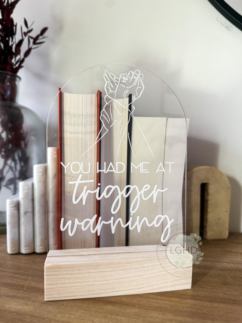 May include: Clear acrylic sign with the text "YOU HAD ME AT trigger warning" in white script. The sign features a line drawing of two hands. The sign is displayed in a wooden base, with books in the background.