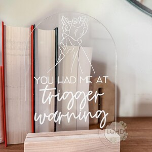May include: Clear acrylic sign with the text "YOU HAD ME AT trigger warning" in white script. The sign features a line drawing of two hands and sits in a light wood base. Books are in the background.