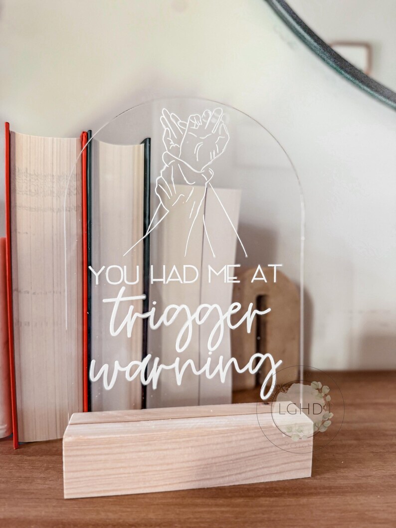 May include: Clear acrylic sign with the text "YOU HAD ME AT trigger warning" in white script. The sign features a line drawing of two hands and sits in a light wood base. Books are in the background.