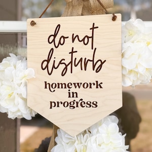 Puede incluir: Un letrero de madera con el texto "No molestar, tarea en progreso" en letras marrones. El letrero está colgado de una cuerda de yute marrón y está rodeado de flores blancas.