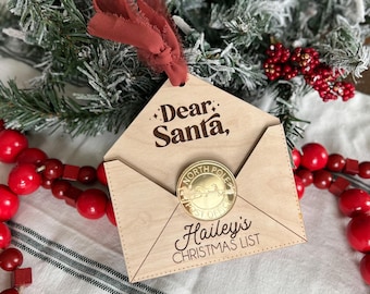May include: A wooden Christmas ornament shaped like an envelope with the text "Dear Santa" on the front. The envelope has a gold circle with the text "North Pole Post Office" and a small image of a sleigh. The envelope also has the text "Hailey's Christmas List" on the front.