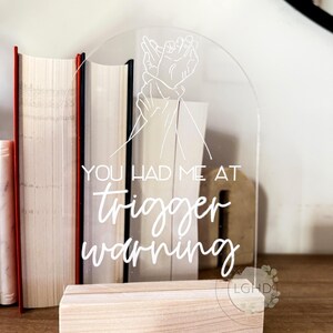 May include: Clear acrylic sign with the text "YOU HAD ME AT trigger warning" in white script. The sign features a line drawing of two hands and rests on a natural wood base. Books are visible in the background.