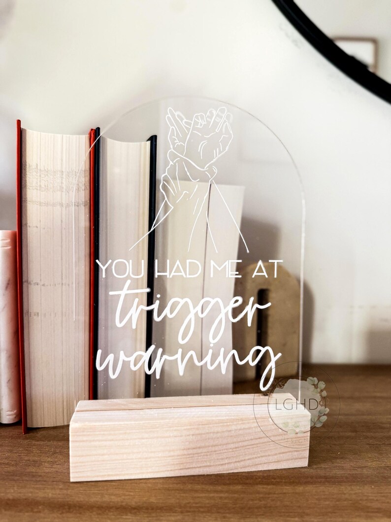 May include: Clear acrylic sign with the text "YOU HAD ME AT trigger warning" in white script. The sign features a line drawing of two hands and rests on a natural wood base. Books are visible in the background.