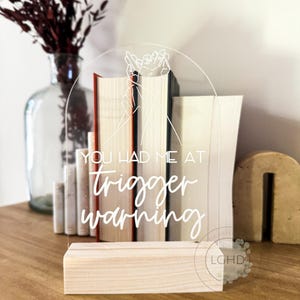 May include: A clear acrylic sign with the text "YOU HAD ME AT trigger warning" in white script. The sign is supported by a light wood base and features a line drawing of hands. Books are visible behind the sign.