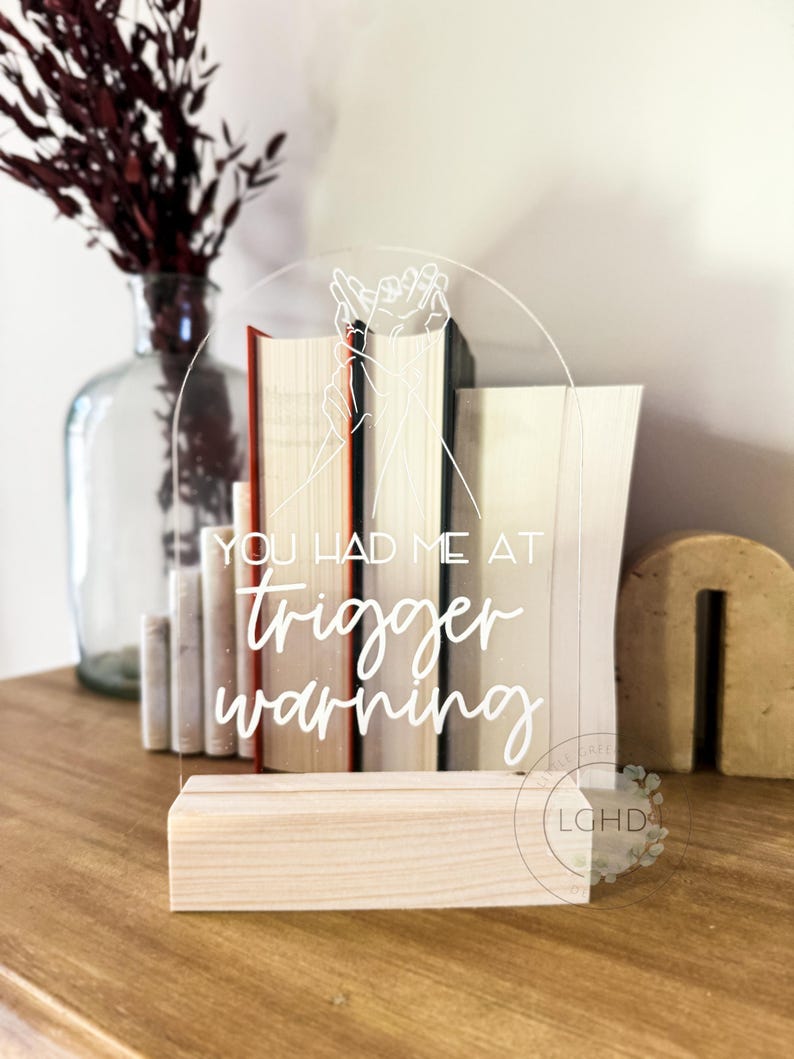 May include: A clear acrylic sign with the text "YOU HAD ME AT trigger warning" in white script. The sign is supported by a light wood base and features a line drawing of hands. Books are visible behind the sign.