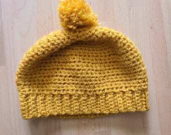 May include: Mustard yellow crochet beanie hat with a matching pom-pom on top. The hat is handmade with a ribbed brim and a textured stitch pattern. This warm hat is perfect for cold weather.