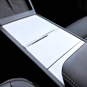 May include: A white center console cover for a vehicle interior featuring a smooth, matte finish with a central horizontal seam and a recessed handle detail, designed to fit over the existing console storage compartment lid.