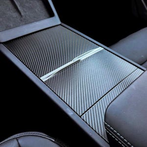 May include: A carbon fiber textured console cover with a silver accent. The cover is black and gray, with a woven pattern. The console cover is installed in a vehicle, with black leather seats visible.