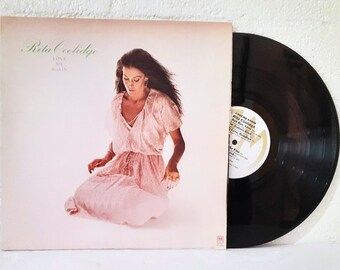 May include: Rita Coolidge album cover featuring a woman in a pink dress sitting on a white background. The album title "Love Me Again" is printed in green text.