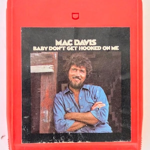 May include: A vintage red 8-track tape of Mac Davis' album, "Baby Don't Get Hooked on Me." The album cover features a photo of Mac Davis in a blue shirt. The tape is in good condition, showing signs of age.