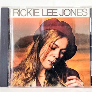 May include: A CD case featuring the album cover for Rickie Lee Jones. The cover art shows a woman with long, flowing hair wearing a burgundy hat, with the artist's name in white block letters at the top.