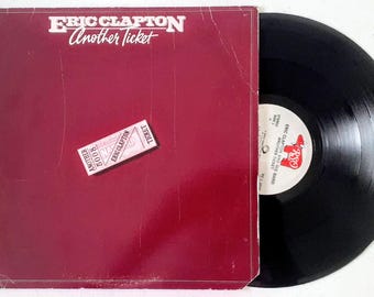 Puede incluir: Una portada de álbum de vinilo rojo con el título "Eric Clapton Another Ticket" y un talón de entrada con el texto "Another Ticket 5008 Eric Clapton" en la parte delantera. La parte posterior de la portada del álbum no es visible. El disco es negro y tiene una etiqueta blanca con el texto "Eric Clapton and His Band Another Ticket" en él.