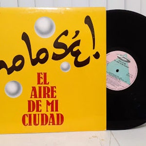 May include: A yellow vinyl record cover with the text "hola sé!" in black and the text "El Aire De Mi Ciudad" in red. The record cover has a black vinyl record on the right side.