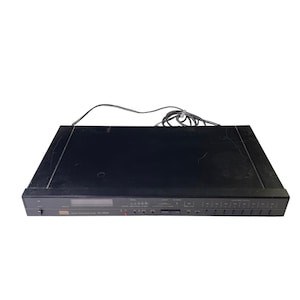 Puede incluir: Sintonizador estéreo negro Sansui TU-300L con un diseño rectangular. El panel frontal tiene una pantalla, botones y perillas. Un cable de alimentación se extiende desde la parte posterior. La unidad está diseñada para recibir señales de radio.