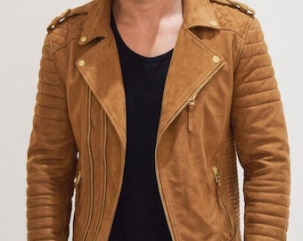 May include: A tan suede motorcycle jacket featuring gold-tone zippers, quilted shoulder and sleeve detailing, a notched lapel, and epaulets. The jacket is worn over a plain black t-shirt. The design includes multiple zippered pockets and cuffs.