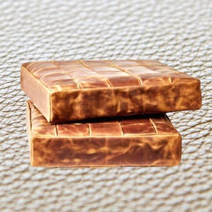 May include: Two brown leather square ottomans stacked on top of each other. The ottomans have a quilted design.