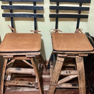Puede incluir: Dos taburetes de barril de madera con cojines de asiento de cuero marrón. Los taburetes tienen un respaldo de metal negro con listones horizontales. Los taburetes están sobre un suelo de madera.