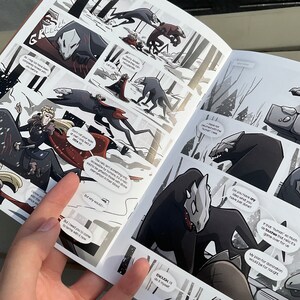 Puede incluir: Páginas de cómic abiertas con ilustraciones en blanco y negro. La obra de arte representa una escena de fantasía con figuras en un bosque nevado. Las burbujas de texto contienen diálogos. El cómic está sostenido por una mano.