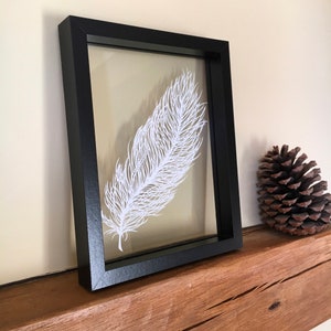 May include: A white paper feather art piece framed in a black frame. The frame is sitting on a wooden surface.