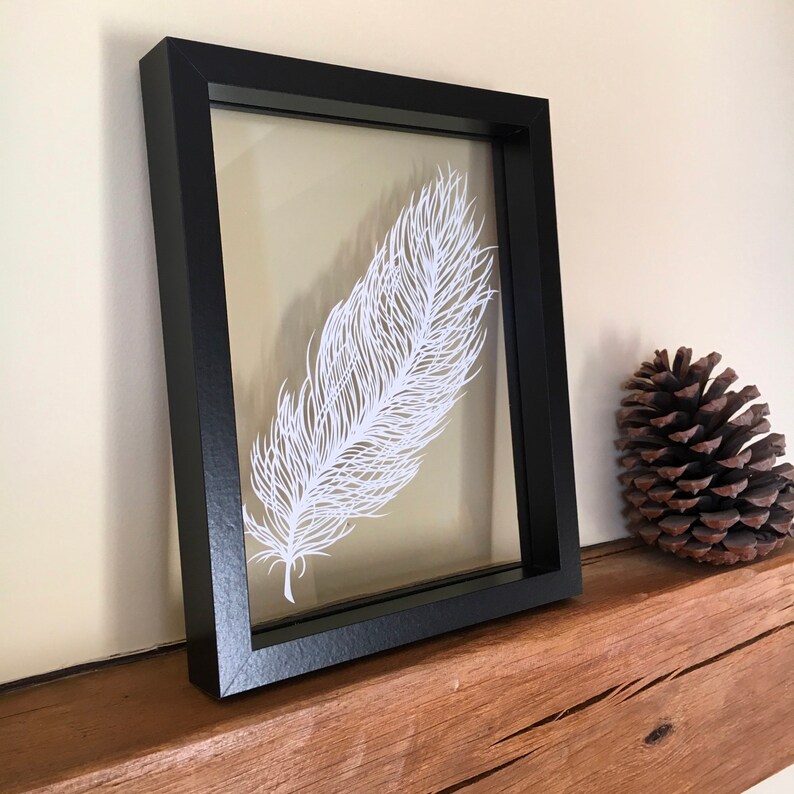 May include: A white paper feather art piece framed in a black frame. The frame is sitting on a wooden surface.