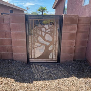 May include: A black metal gate with a tree design cut out. The tree is made of a copper-colored metal. The gate is set in a brick wall.