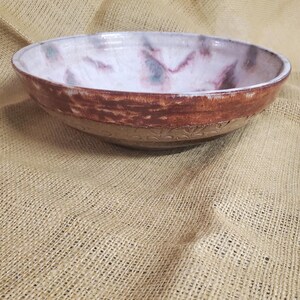May include: A large, white ceramic bowl with a brown rim and a decorative floral pattern on the bottom. The bowl has a mottled, marbled glaze with shades of pink, purple, and green.