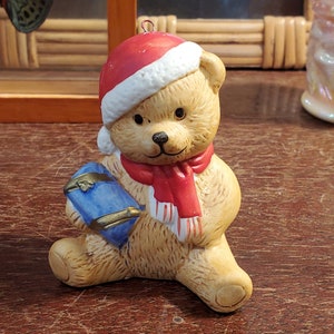 May include: A ceramic Christmas ornament of a teddy bear wearing a red Santa hat with white trim and a red scarf with white stripes. The bear is holding a blue gift box.