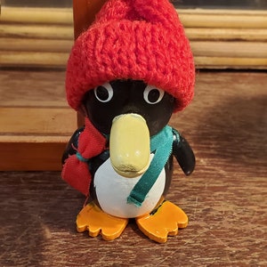 May include: A wooden penguin figurine wearing a red knitted hat with a white pom pom and a green scarf. The penguin has a yellow beak and orange feet.