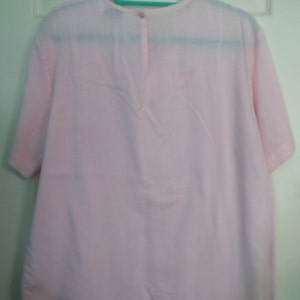 Op de afbeelding: Een lichtroze blouse met korte mouwen en een knoopsluiting aan de achterkant van de hals.