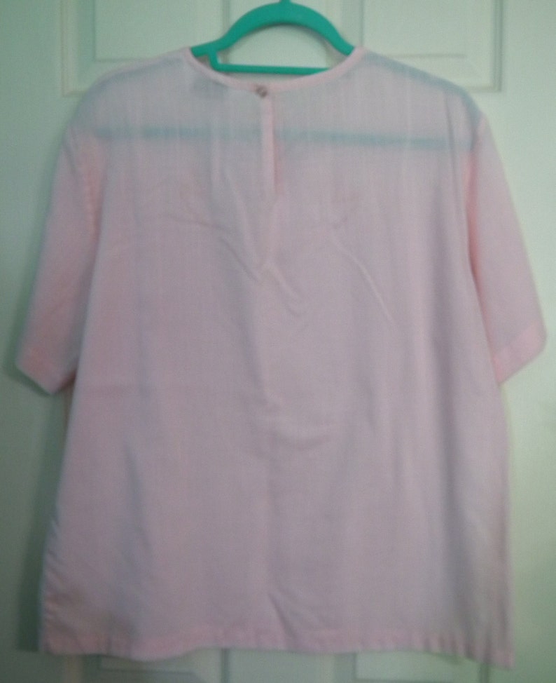 Op de afbeelding: Een lichtroze blouse met korte mouwen en een knoopsluiting aan de achterkant van de hals.