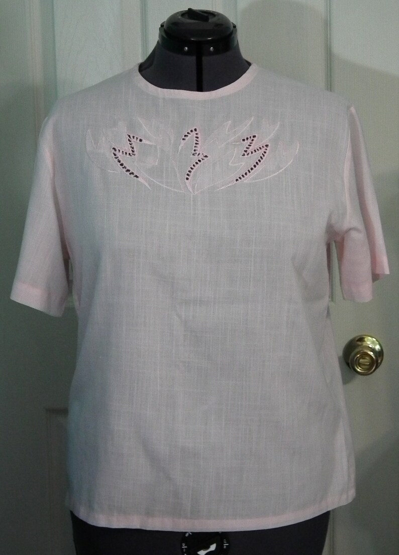 Op de afbeelding: Een lichtroze blouse met korte mouwen en een delicaat geborduurd ontwerp op de halslijn. De borduurwerk heeft een bloemmotief met een zwarte omtrek.
