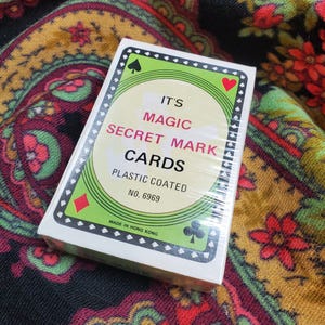 Op de afbeelding: Een rechthoekig kaartspel in een witte doos met een groene rand. In het midden staat de tekst IT'S MAGIC SECRET MARK CARDS, PLASTIC COATED, NO. 6969 en MADE IN HONG KONG. De rand toont zwarte kaartkleursymbolen.