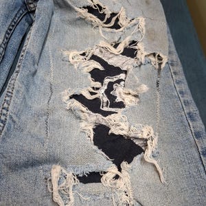 May include: Light blue denim jeans featuring a large distressed section on the thigh. The area has multiple horizontal rips with frayed white edges, revealing a dark navy blue fabric patch underneath that is secured with visible zigzag stitching.
