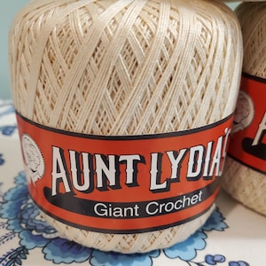 Könnte beinhalten: Nahaufnahme mehrerer Spulen Aunt Lydia's Giant Crochet-Garn. Das Garn ist hellcremefarben und fest zu einer Kugel gewickelt. Das Etikett ist orange mit schwarzem Text "AUNT LYDIA'S Giant Crochet".