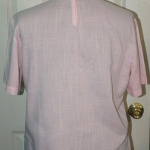 Op de afbeelding: Een lichtroze blouse met korte mouwen en een knoopsluiting op de rug.