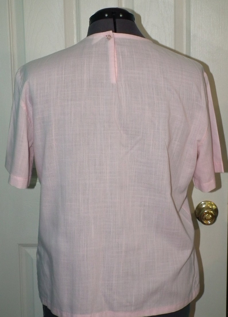 Op de afbeelding: Een lichtroze blouse met korte mouwen en een knoopsluiting op de rug.