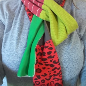 May include: A colorful scarf with stripes, leopard print, and solid green and lime green fabric. The scarf is tied around the neck of a person wearing a gray sweater.