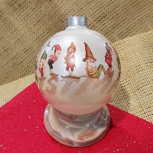 Könnte beinhalten: Weißer gläserner Weihnachtsbaumschmuck mit einer Darstellung von sieben Zwergen in verschiedenen Posen. Der Schmuck befindet sich auf einem Ständer mit Marmormuster.