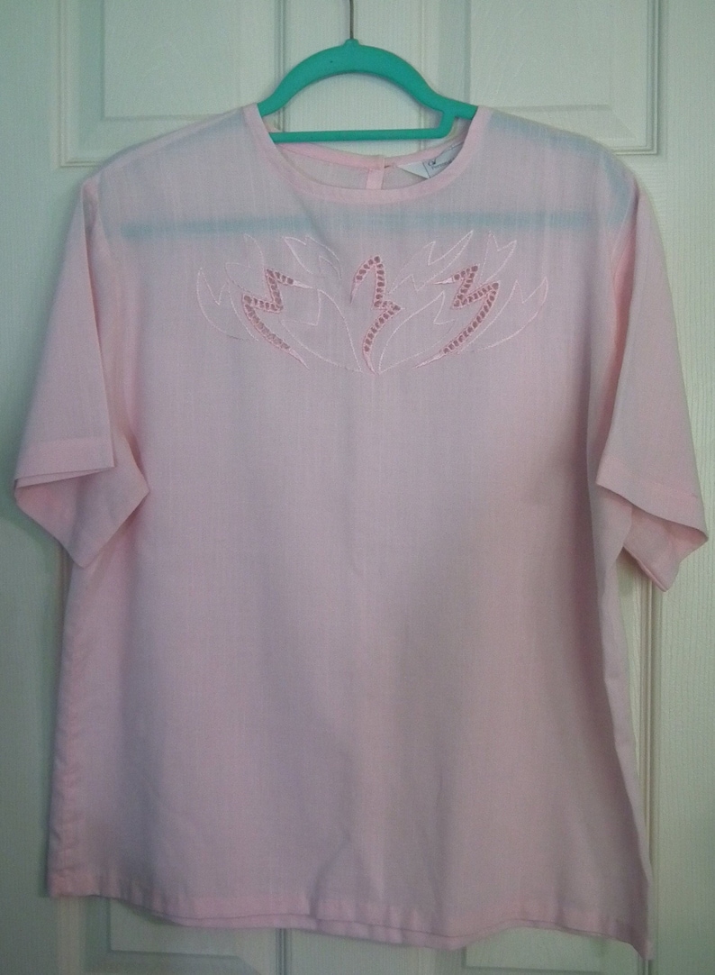 Op de afbeelding: Een roze blouse met korte mouwen en een wit geborduurd ontwerp van gestileerde bladeren aan de voorkant.