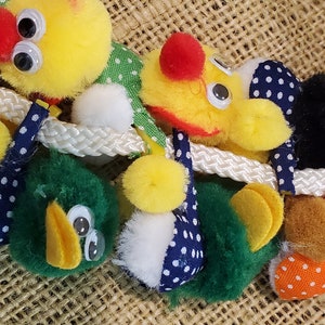 May include: A set of colorful plush toys with googly eyes and polka dot fabric. The toys are attached to a white rope and appear to be hanging from a burlap background.