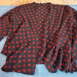 May include: A long-sleeved open-front cardigan featuring a black base colour with a repeating pattern of red polka dots. The garment has a relaxed, flowing silhouette with a straight hemline and long sleeves, crafted from a lightweight, semi-sheer fabric.