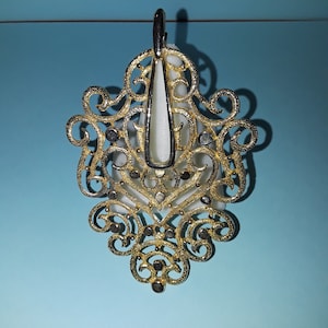 Op de afbeelding: Een gouden filigraan hanger met een wit centraal stuk. De hanger is versierd en gedetailleerd, met een delicaat ontwerp.