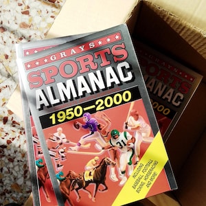 Può includere: Un libro Grays Sports Almanac 1950-2000 con copertina argentata, testo rosso e stelle. La copertina mostra illustrazioni di atleti in vari sport, tra cui football, hockey e corse di cavalli, con un banner giallo nell'angolo.
