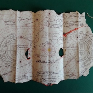 Può includere: Una mappa in stile vintage con un aspetto invecchiato, con linee intricate, testo e un design a rosa dei venti. La mappa ha un bordo strappato e diversi fori, con segni rossi e testo in spagnolo. Un filo è attaccato.