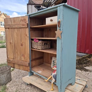 Puede incluir: Un armario de madera pintado de azul con una puerta abierta, que cuenta con estantes interiores de madera natural, una pequeña cesta de mimbre, un adorno de estrella de madera colgado en el lateral y una figura de gallo en el estante inferior.