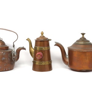 May include: Three antique copper teapots with brass accents. The teapot on the left has a rounded body and a hinged lid. The middle teapot has a tall, cylindrical body with a brass band around the middle. The teapot on the right has a rounded body with a brass lid and a wooden handle.