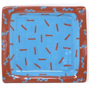 May include: Rectangular ceramic serving tray with a light blue interior and a brown border. The interior features scattered orange dashes. The brown border is decorated with blue squiggles and orange dots. The tray has rounded corners.